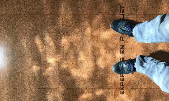 Pintura para corcho con un diseño de baldosas y sombras de luz. Se ven unos pies con zapatos oscuros y pantalones vaqueros.