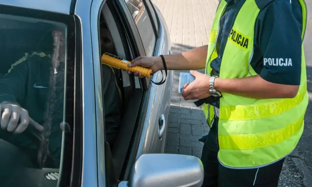 Policjant bada kierowcę alkomatem, sprawdzając, czy nie ma **alkoholu w organizmie**. Kierowca siedzi za kierownicą, a funkcjonariusz trzyma urządzenie przy jego twarzy.