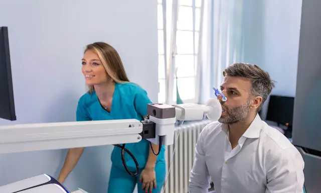 Mann macht Lungenfunktionstest, Arztin überwacht. Ergebnisse zeigen 70 Prozent Lungenfunktion.