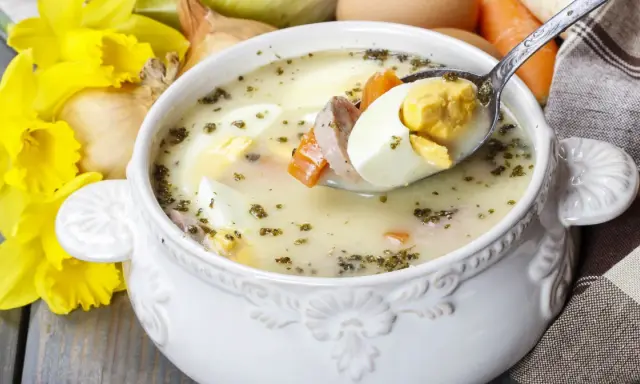 Aromaticzny żur z jajkiem i kiełbasą, podany w ozdobnej misce, z żółtymi narcyzami w tle.