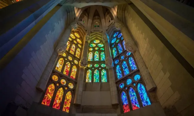 Ventanas de vidrieras de colores vibrantes, que narran la historia del vidrio, iluminan el interior de una catedral con tonos cálidos y fríos.