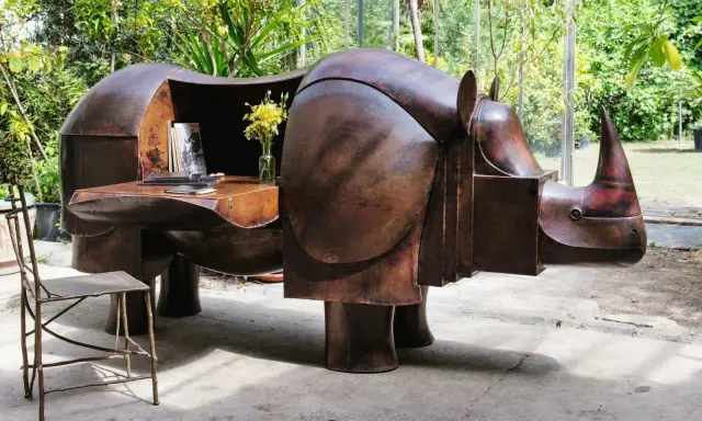Étonnante sculpture lalanne en forme de rhinocéros, bureau fonctionnel en cuivre patiné, avec un vase de fleurs jaunes et une chaise en métal.