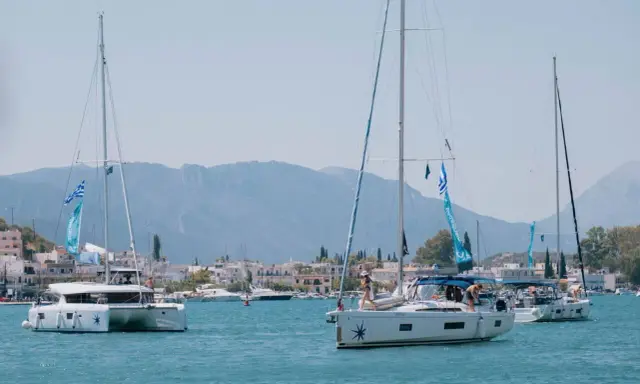 Trois voiliers naviguent dans une baie ensoleillée. Au premier plan, un catamaran monocoque blanc avec des voiles bleues et blanches. Derrière, un autre voilier blanc avec des personnes à bord. Au loin, une ville côtière et des montagnes.