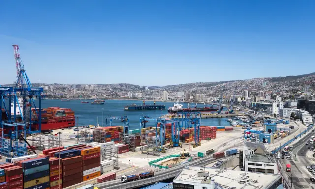 Un vasto porto con gru, container colorati e navi cargo, con una città collinare sullo sfondo. Sembra il porto più grande del mondo.