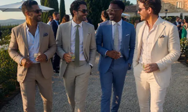 Four stylish men in suits and chinos walk together at a wedding, holding drinks and smiling.