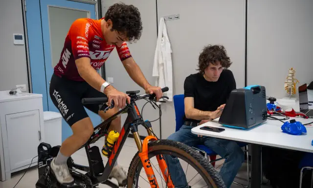 Ciclista su rulli monitorato per l'acido lattico, con un tecnico che osserva i dati su un dispositivo.