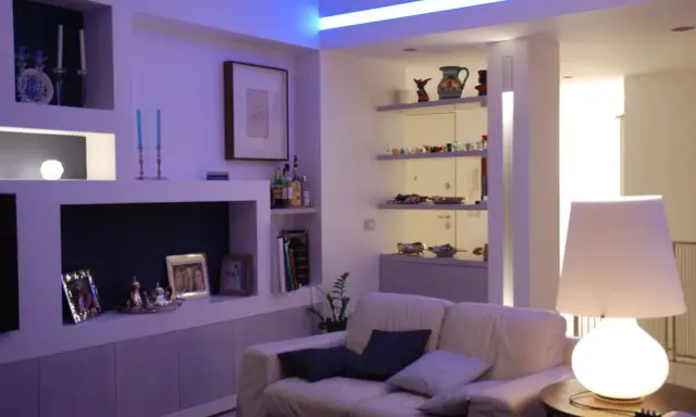 Soggiorno moderno con illuminazione soffusa, ideale per come illuminare un soggiorno con poca luce. Divano bianco, mensole con oggetti e lampada da tavolo.