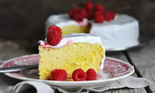 Wilgotne ciasto tres leches z malinami na talerzyku, w tle cały tort.