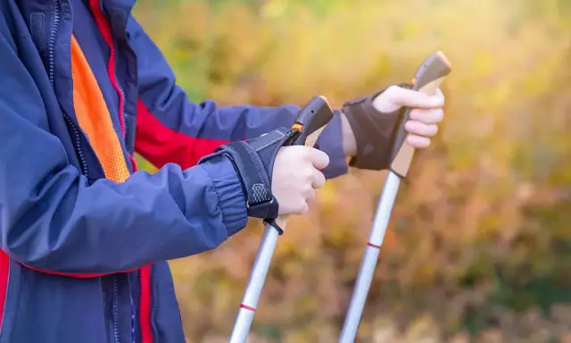Osoba w kurtce ćwiczy, jak chodzić z kijkami trekkingowymi. Dłonie pewnie trzymają uchwyty, gotowe do marszu.
