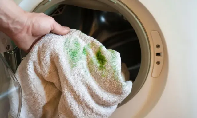 Hand hält Handtuch mit grünen, eingewaschenen Flecken vor einer Waschmaschine.