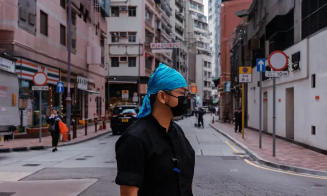 Uomo con bandana blu e mascherina nera in una foto di strada con edifici alti e traffico.