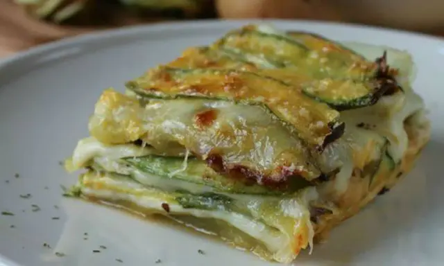 Una fetta di parmigiana di zucchine, stratificata con formaggio filante e zucchine grigliate, servita su un piatto bianco.