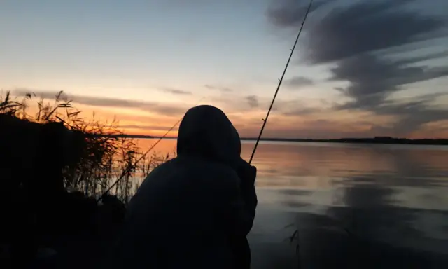 Zachód słońca nad jeziorem, wędkarz czeka na branie. Zastanawia się, jaka będzie karta wędkarska cena.