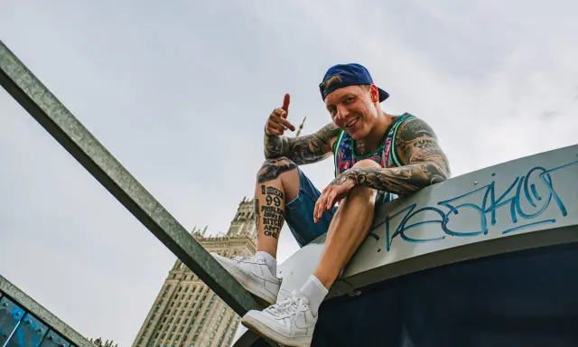 Uśmiechnięty raper z tatuażami, w czapce i kolorowej koszulce, siedzi na dachu, promując swój mixtape "sos ciuchy i borciuchy".