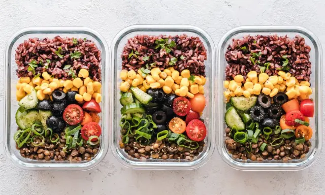 Drei Glasbehälter mit einem gesunden Essensplan für Fitness: Linsen, Gurken, Oliven, Tomaten, Mais und brauner Reis.