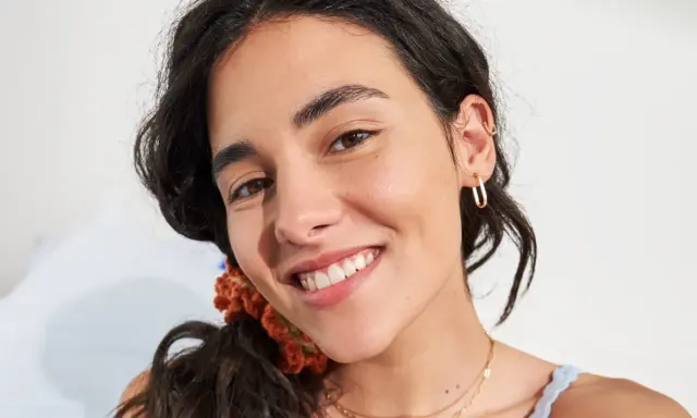 Primo piano di una giovane donna con un sorriso radioso, la sua fronte lucida riflette la luce. Indossa orecchini a cerchio dorati e una collana sottile.