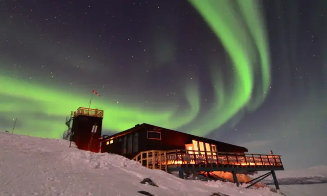 Norrsken dansar över en snöig nationalpark i Lappland. En byggnad med upplysta fönster och en tornliknande struktur står på en sluttning.