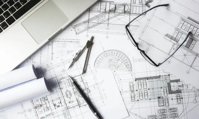Na stole architekta leżą plany budynku, laptop, okulary i cyrkiel. To miejsce pracy nad projektem i jego kosztorysem.