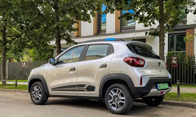 Srebrny Dacia Spring, elektryczny samochód miejski, parkuje pod drzewami. Jego zasięg pozwala na swobodne poruszanie się po mieście.