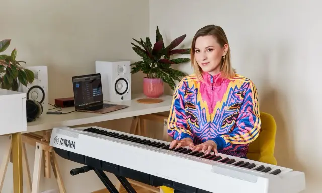 Kobieta gra na pianinie Yamaha. W tle laptop i głośniki, idealne do nauki śpiewu dla dzieci.