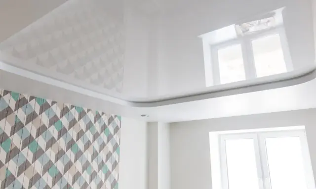 Biały, błyszczący sufit napinany z subtelnymi wzorami geometrycznymi, odbijający światło okna.