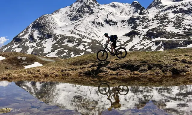 Mountainbiker auf einem epischen Alpencross-Abenteuer, die schneebedeckten Gipfel spiegeln sich in einem Bergsee.