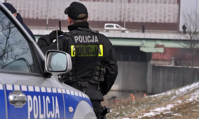 Policjant w kamizelce z napisem "POLICJA", zgodnej z rozporządzeniem w sprawie umundurowania policjantów, stoi obok radiowozu.