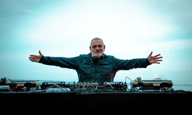 DJ Ralf, un vero patrimonio della musica elettronica, con le braccia aperte davanti alla sua consolle, pronto a far ballare il mondo.