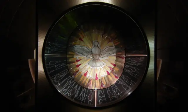 Symbol Ducha Świętego w witrażu, otoczony promieniami światła i symbolizujący siedem darów ducha świętego.