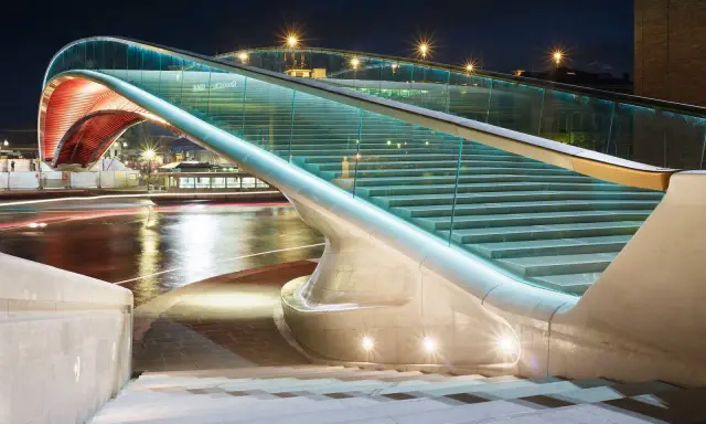 Le pont de la Constitution, illuminé la nuit, avec ses escaliers de verre et ses arches rouges, surplombe un canal aux reflets lumineux.