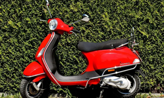 Scooter rojo brillante aparcado frente a un seto verde. ¡Listo para salir con mi permiso AM!