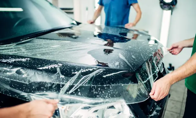 Profesjonaliści nakładają folię ochronną na maskę czarnego auta. To kluczowy etap car wrappingu, chroniący lakier.