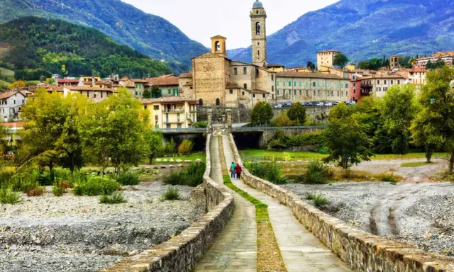 Ponte Gobbo e borghi vicino a Bobbio, con montagne sullo sfondo. Due persone passeggiano sul ponte antico.