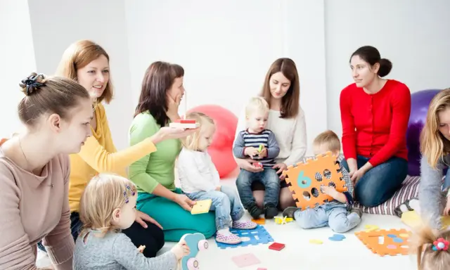 Grupa dzieci i dorosłych uczestniczy w zajęciach, które przypominają terapię pedagogiczną. Dzieci bawią się klockami i zabawkami.