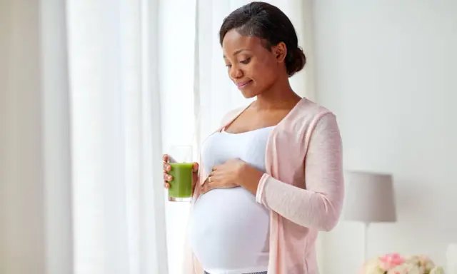 Femme enceinte tenant un verre de jus vert, caressant son ventre. Elle prend soin de sa peau sèche pendant la grossesse.