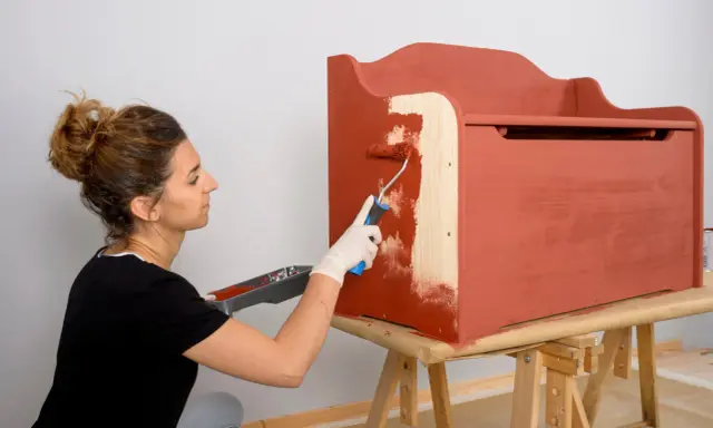 Mujer con guantes blancos pinta con rodillo una caja de madera de color terracota.