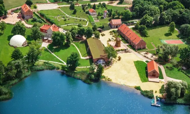 Widok z lotu ptaka na ośrodek SPA nad jeziorem z budynkami, plażą i kortem tenisowym.