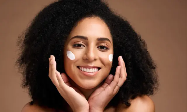 Une femme souriante applique du beurre de karité visage sur ses joues. Ses mains caressent doucement sa peau.