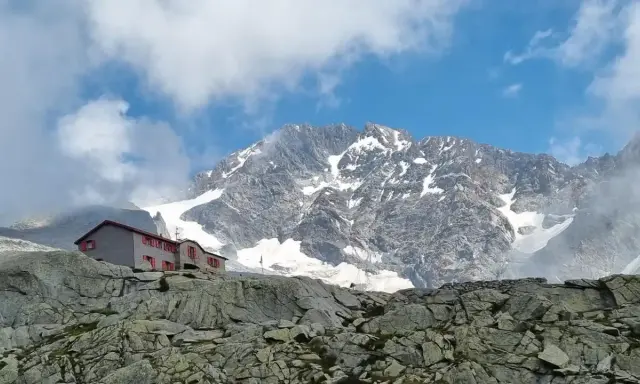 Rifugio Gherardi, un edificio con finestre rosse, si erge su un pendio roccioso sotto un imponente massiccio montuoso innevato.