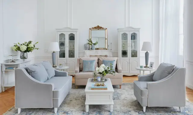 Salotto in stile shabby chic con divani grigi, poltrone, tavolino bianco, specchio dorato e vetrine bianche.