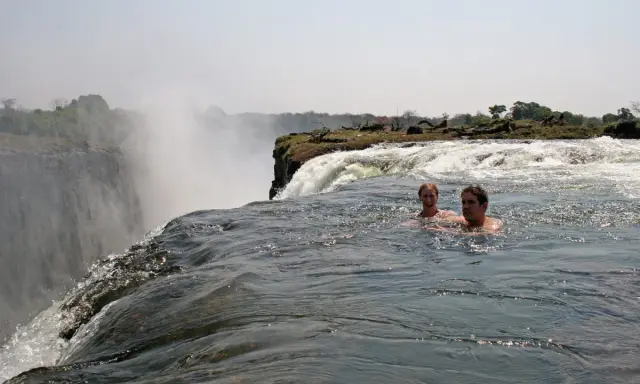 Zwei Personen schwimmen im Devils Pool am Rande der Victoriafälle, mit Gischt und üppiger Vegetation im Hintergrund.