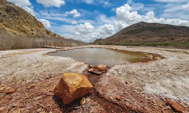 Un estanque natural en Les Salinetes de Novelda, rodeado de rocas y vegetación seca, bajo un cielo azul con nubes blancas.