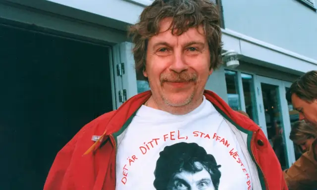 Staffan Westerberg, känd för sina filmer och tv-program, ler i en röd jacka över en t-shirt med hans eget ansikte och texten "Det är ditt fel, Staffan Westerberg".