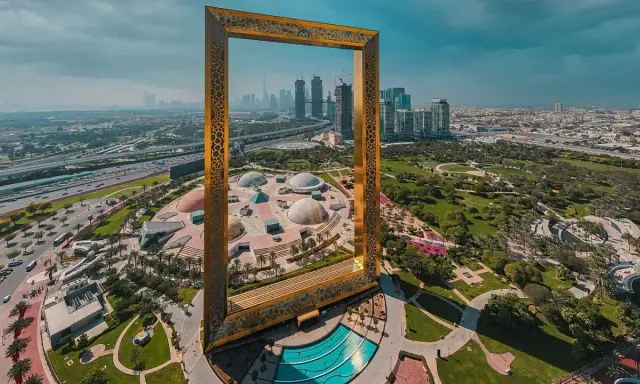 Dubai Frame rahmt die Skyline und die futuristischen Dubai Projekte ein. Ein Park mit Kuppeln und ein Pool sind im Vordergrund.