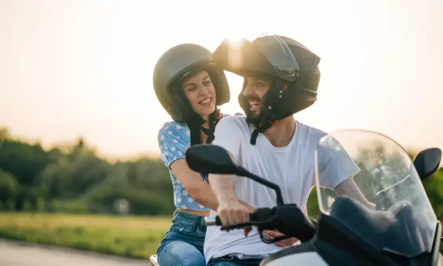 Pareja sonriente en moto, ella con blusa floral y él con camiseta blanca. Listos para disfrutar de la carretera con su carnet A2.