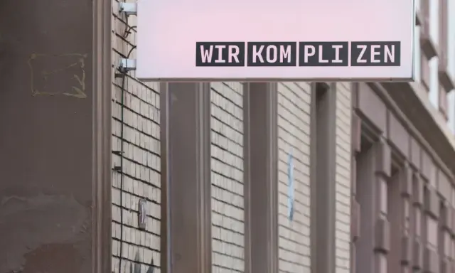 Leuchtreklame mit dem Schriftzug "WIR KOMPLIZEN" über einer Fassade.