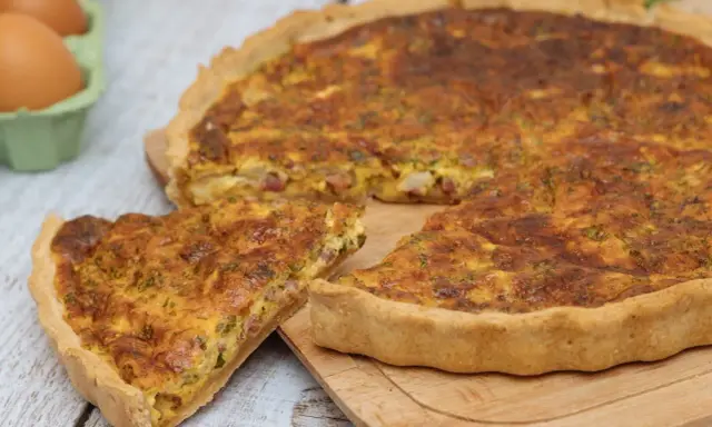 Une part de quiche lorraine, plat originaire de Lorraine, dorée et appétissante, sur une planche en bois. Des œufs frais sont visibles en arrière-plan.