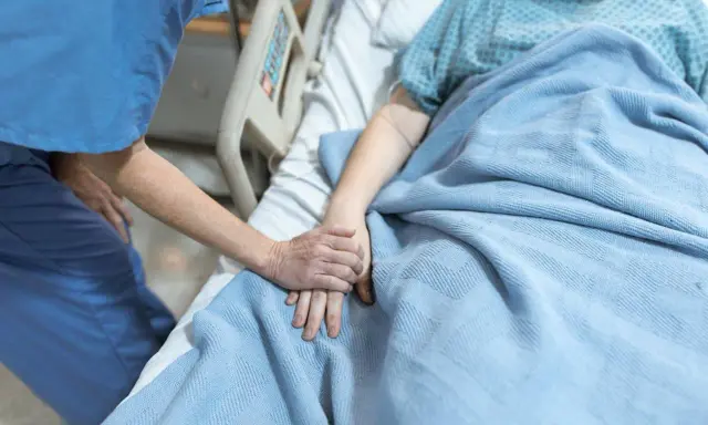 Pielęgniarka trzyma za rękę pacjentkę leżącą w łóżku szpitalnym pod niebieskim kocem.