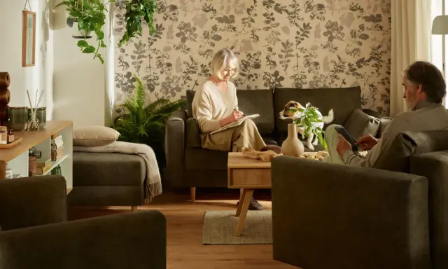 Przytulny salon, gdzie kobieta pisze, a mężczyzna korzysta z tabletu. Idealne inspiracje, jak urządzić pokój dla seniora, z wygodnymi meblami i roślinnością.