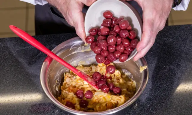 Hände geben Kirschen in eine Schüssel mit Teig für ein einfaches Kirschenmichel Rezept. Ein roter Teigschaber liegt daneben.